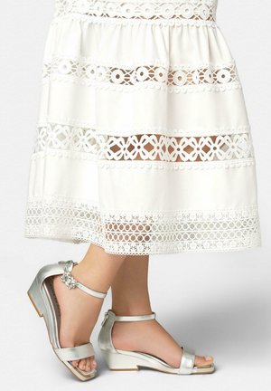 Stopy w srebrnych, niskich sandałach na klinowym obcasie z paskami wokół kostki oraz biała koronkowa spódnica midi z misternymi kwiatowymi wzorami.