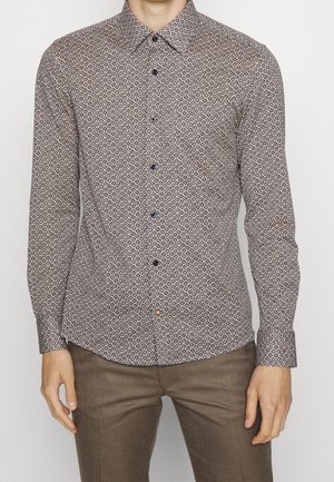 Mann trägt ein eng anliegendes Langarmhemd mit kleinem Rautenmuster und braune Hose, steht vor einem schlichten Hintergrund.