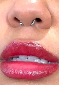 Nærbilde av lepper med glansfull, lys rød leppestift, som har synlige hvite tenner og en sølv septumpiercing i neseborene.