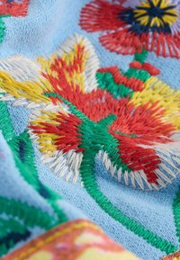 Kleurrijke bloemenborduurwerk met rode, gele, groene en witte draden op zacht lichtblauw stof met gestructureerde stikdetails.
