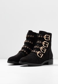 Botines de ante negro con cuatro tiras de hebillas doradas y cremallera lateral sobre una superficie blanca y fondo gris.
