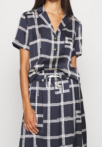 Vestido camisero azul marino con estampado y mangas cortas, que presenta líneas de cuadrícula blancas y acentos de texto, un frente abotonado y una cintura con cordón.