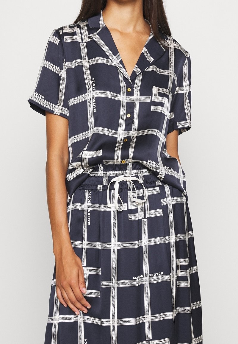 Abito camicia blu navy stampato con maniche corte, caratterizzato da linee bianche a griglia e accenti di testo, una chiusura frontale con bottoni e una vita regolabile con coulisse.