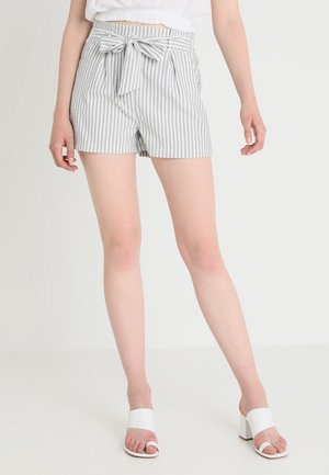 Femme portant un short gris et blanc à rayures verticales taille haute avec ceinture nouée, sandales blanches à talons et un top blanc court.