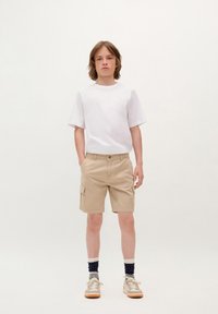 Maglietta bianca a maniche corte, pantaloni cargo beige chiaro con tasche laterali, abbinati a sneaker beige e calzini con fantasia. Outfit semplice e casual.