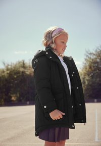 Čierna prešívaná bunda s kapucňou, vybavená cvočkami a bočnými vreckami. Dieťa má na sebe biele tričko a plisovanú sukňu. Prírodná scenéria.