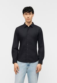 Νεαρός άνδρας που κοιτάζει μπροστά, φορώντας εφαρμοστό μαύρο πουκάμισο με κουμπιά και ανοιχτόχρωμο μπλε τζιν, σε απλό λευκό φόντο.