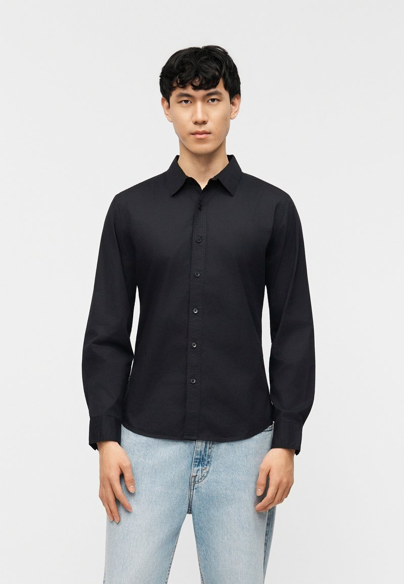 Νεαρός άνδρας που κοιτάζει μπροστά, φορώντας εφαρμοστό μαύρο πουκάμισο με κουμπιά και ανοιχτόχρωμο μπλε τζιν, σε απλό λευκό φόντο.