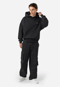 Sort hættejakke med en frontlomme og logo, parret med sorte cargo-bukser med store sidelommer. Modellen har hvide sneakers på.