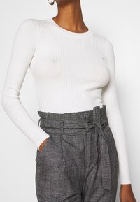 Weißes, geripptes Langarmoberteil kombiniert mit hochtaillierten grauen Karo-Hosen, die einen gebundenen Gürtel besitzen. Beide Teile haben eine glatte Textur.