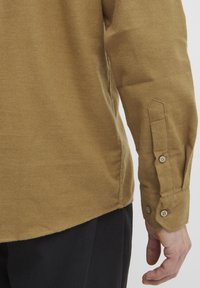 Cămașă lungă maro cu mâneci lungi, guler cu nasture și slit lateral. Țesătura pare texturată și ușor periată, oferind un aspect casual.