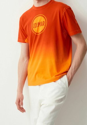 Persoon draagt een oranje gradient T-shirt met het "COLMAR" logo en witte broek, staand tegen een effen lichte achtergrond.