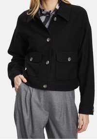 Žena v černé košilové bundě s klopovými kapsami, pod ní šedý vzorovaný top a šedé skládací kalhoty, ruka v kapse.