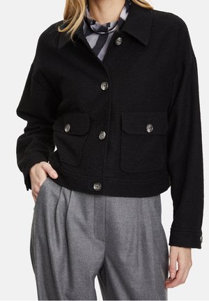 Kvinna som bär en svart knappjacka med lockfickor över en gråmönstrad topp och grå veckade byxor, med handen i fickan.