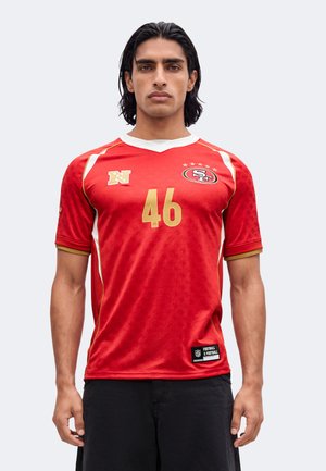 Junger Mann mit dunklen Haaren trägt ein rotes Sporttrikot mit goldener Nummer 46 und schwarze Hose, steht vor einem schlichten hellen Hintergrund.