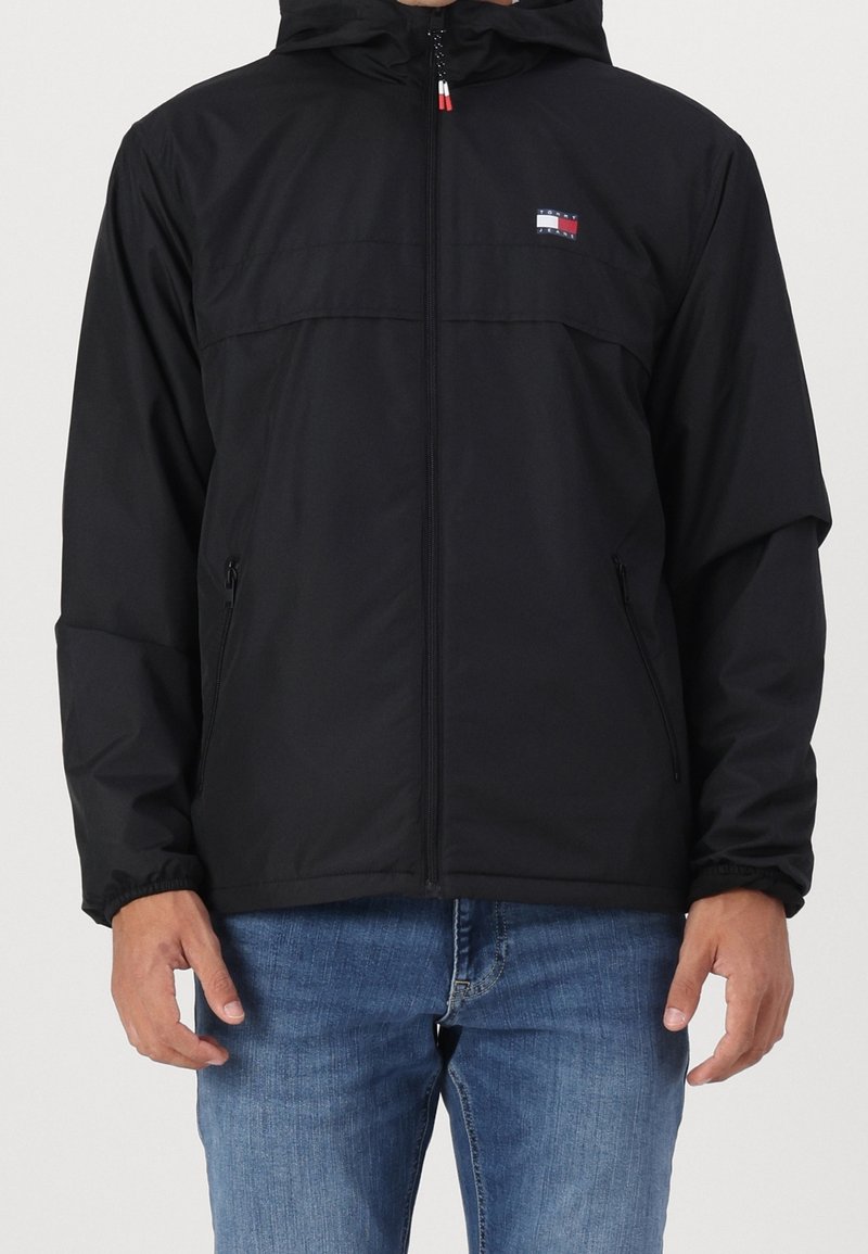 Schwarze Kapuzenjacke mit Reißverschluss vorne, elastischen Bündchen, Seitentaschen und einem Logodetail. Leicht, glatte Textur, lässige Passform.