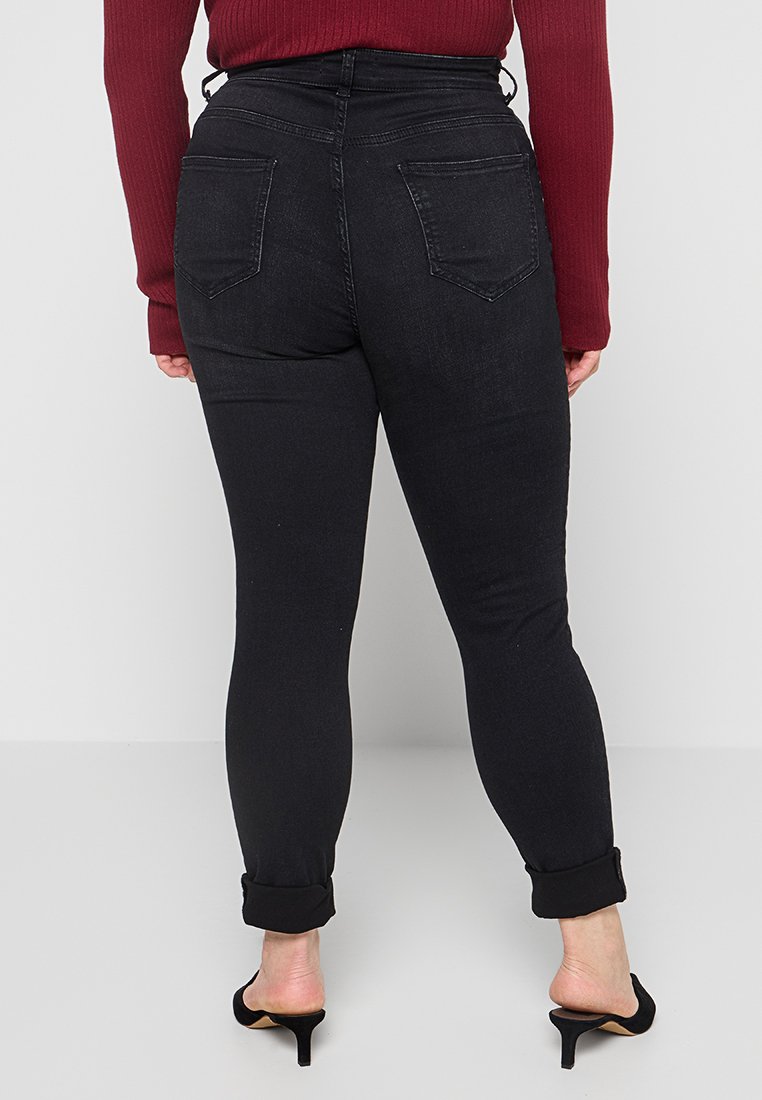 Schwarze hochgeschnittene Skinny-Jeans mit zwei Gesäßtaschen, umgeschlagenen Säumen und einer glatten Textur, kombiniert mit einem gerippten burgunderfarbenen Oberteil.