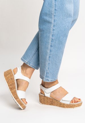 Feet wearing white platform sandals with cork wedge heels and light blue jeans against a plain background.