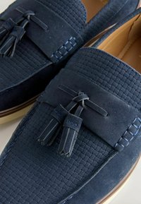 Paire de mocassins en daim bleu marine avec texture tissée et accents de pompons à l'avant, dotés de semelles intérieures en cuir camel et de semelles marron.