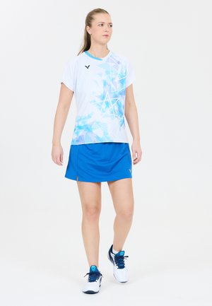 Kvinde gående iført hvid og blå mønstret sportstrøje, blå atletisk nederdel og hvide sneakers på ensartet hvid baggrund.