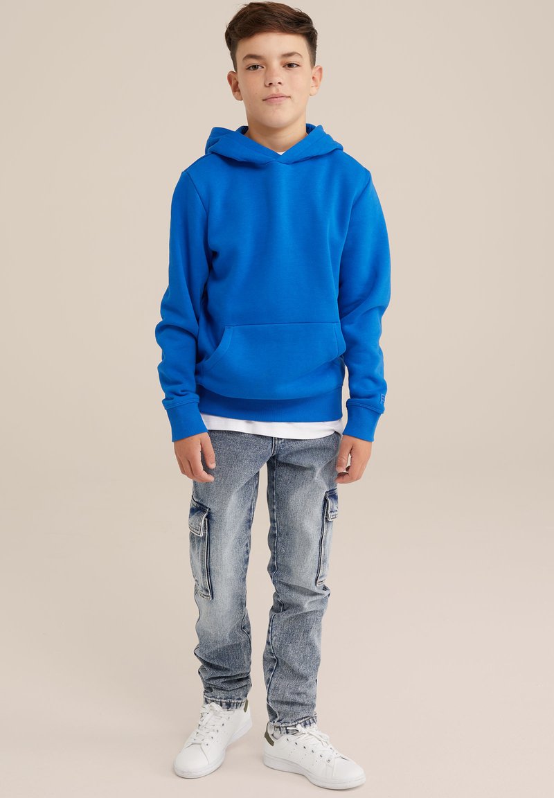 Blaue Kapuzenjacke aus Baumwollmischung mit einer Fronttasche. Kombiniert mit hellblauen Cargo-Jeans und weißen Sneakern.