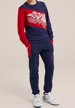 Dziecko w granatowo-czerwonym dresie z napisem "BAD BOYS" na bluzie i białymi trampkami, z rękami w kieszeniach, stojące na neutralnym tle.