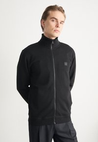 Black zip-up sweatshirt with a high collar, made of soft fabric. Features a small logo patch on the left chest and zippered front.