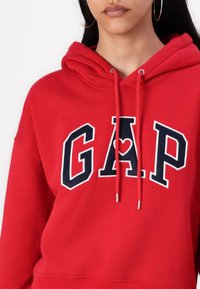 Czerwona bluza z kapturem z granatowym logo "GAP", białymi akcentami i srebrnymi końcówkami sznurków. Miękka, przyjemna w dotyku tkanina.
