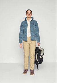 Blaue gemusterte Jacke, beiges Hemd, beige Hose und braune Wanderschuhe. Schwarzer und grauer Rucksack in der Hand gehalten. Einfacher Hintergrund.
