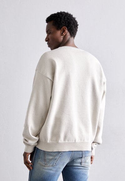 Pull beige clair avec un ourlet et des poignets côtelés, présentant une coupe décontractée et un col rond, porté avec un jean bleu.
