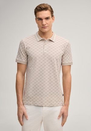 Młody mężczyzna ubrany w beżową, wzorzystą koszulkę polo z krótkim rękawem i kołnierzykiem oraz jasne spodnie, stojący na jednolitym tle.