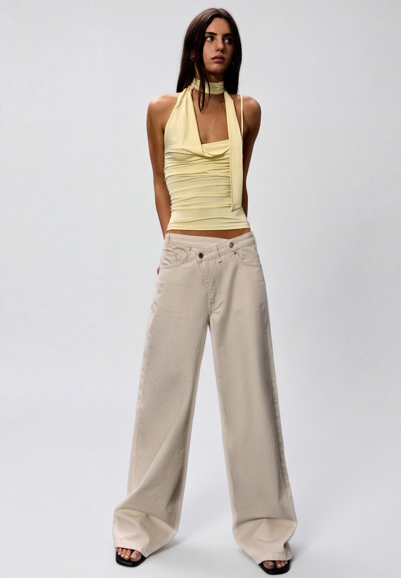 Femme portant un haut dos nu plissé jaune pâle avec un lien au cou et un pantalon beige ample à jambes larges, debout devant un fond clair uni.