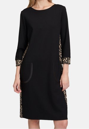 Kobieta ubrana w czarną sukienkę do kolan z rękawami 3/4, bocznymi panelami w panterkę, mankietami i przednimi kieszeniami naszywanymi.