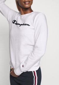 Kvinna iförd en vit Champion sweatshirt med svart logotyp, svarta byxor med röd, vit och blå sidränder, poserar mot en ljus bakgrund.