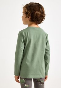 Langærmet grøn bomuldsskjorte med rund hals og glat tekstur, båret med grå leggings med farverige dyreprint.