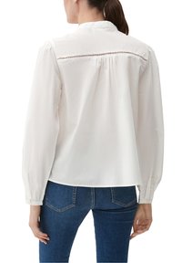Donna con una camicia bianca a maniche lunghe con dettaglio nella parte posteriore e jeans in denim blu, mostrata da dietro su uno sfondo bianco.