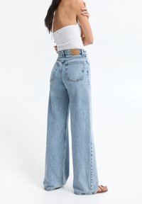 Jean large en denim bleu clair à taille haute, avec poches arrière et patch en cuir à la ceinture, associé à un haut blanc.