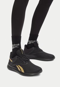 Čierne a zlaté tenisky Reebok nosené s čiernymi ponožkami Reebok a čiernymi legínami, zobrazené od strednej časti lýtok po nohy na bielom pozadí.