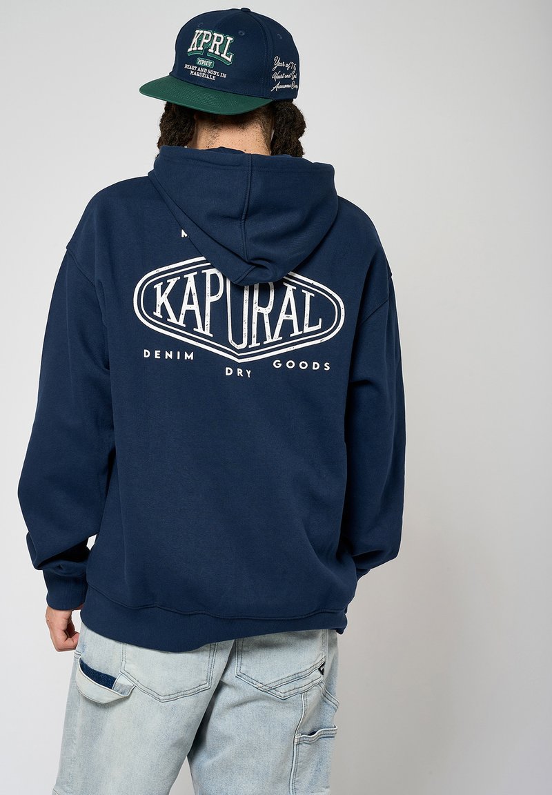 Sweat à capuche bleu marine avec le texte blanc "KAPURAL DENIM DRY GOODS" au dos. Matière en mélange de coton avec une poche kangourou et une capuche réglable.