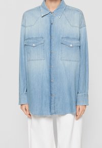 Světle modrá džínová košile na knoflíky s dvěma kapsami na hrudi, nošená přes bílé kalhoty, zobrazená od krku po střed stehna na stojící osobě.