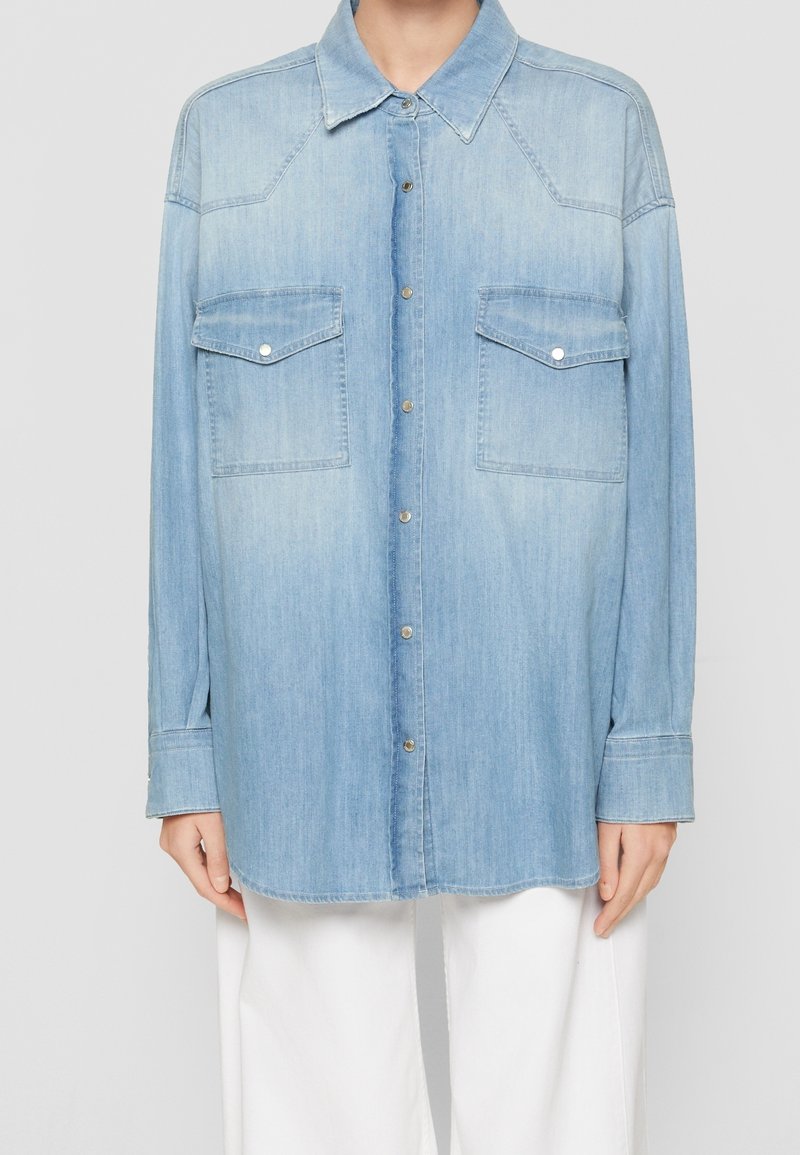 Světle modrá džínová košile na knoflíky s dvěma kapsami na hrudi, nošená přes bílé kalhoty, zobrazená od krku po střed stehna na stojící osobě.