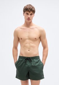 Πράσινο μαγιό με ελαστική ζώνη μέσης και μαύρο κορδόνι, με πλαϊνές τσέπες και μικρή λεπτομέρεια με λογότυπο στο κάτω μέρος.