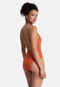 Maillot de bain une pièce orange, présentant un motif en zigzag, un dos décolleté et de fines bretelles spaghetti, mettant en valeur un tissu lisse et texturé.