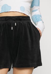 Black velour shorts featuring an elastic waistband, drawstring with metal tips, and side pockets. Smooth texture and relaxed fit.