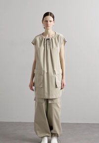 Tunika beige con scollatura arricciata, maniche corte e due tasche frontali, abbinata a pantaloni beige larghi e scarpe bianche.