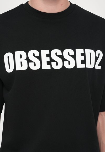 T-shirt noir à manches courtes avec un texte blanc en gras "OBSESSED2" sur la poitrine, fabriqué en tissu en coton lisse.