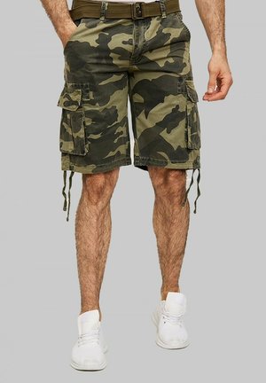 Shorts cargo camouflés dans des tons de vert et de marron, dotés de poches latérales, d'un cordon de serrage et de passants de ceinture. Associés à des baskets blanches.
