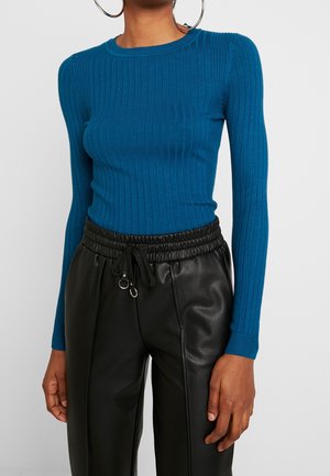 Femme portant un pull bleu côtelé à manches longues et un pantalon en cuir noir taille haute avec une ceinture à cordon et des boucles d'oreilles anneaux.