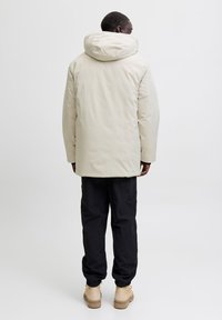 Mees, kes kannab beeži kapuutsiga talvejope, musti pükse ja beeže saapaid, seisab valgel taustal selg vaataja poole.