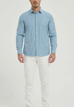 Homme portant une chemise à manches longues rayée verticalement bleu et blanc avec un pantalon blanc, sur un fond uni.